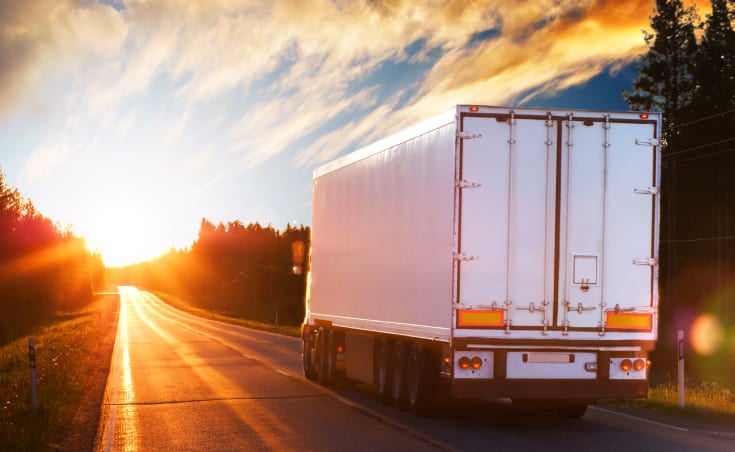 A white semi-truck hauls a load down an open road as the sun sets.