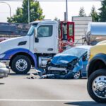 A semi-truck sits in the foreground of the picture at a traffic light next to a totaled blue car.